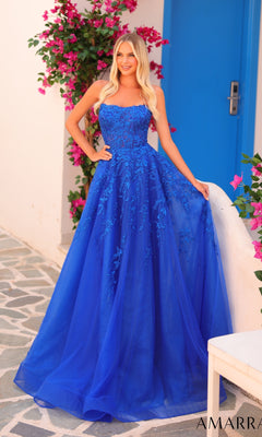 A woman in the Amarra 88258 long prom dress, bright blue with floral lace, stands by a white wall and blue door, surrounded by pink flowers.