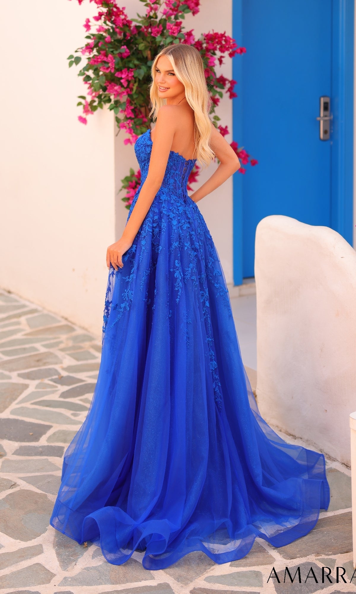 A woman wearing the Amarra 88258 long prom dress stands outdoors on a stone path next to white walls, pink flowers, and a blue door.