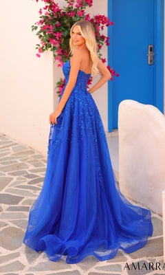 A woman wearing the Amarra 88258 long prom dress stands outdoors on a stone path next to white walls, pink flowers, and a blue door.