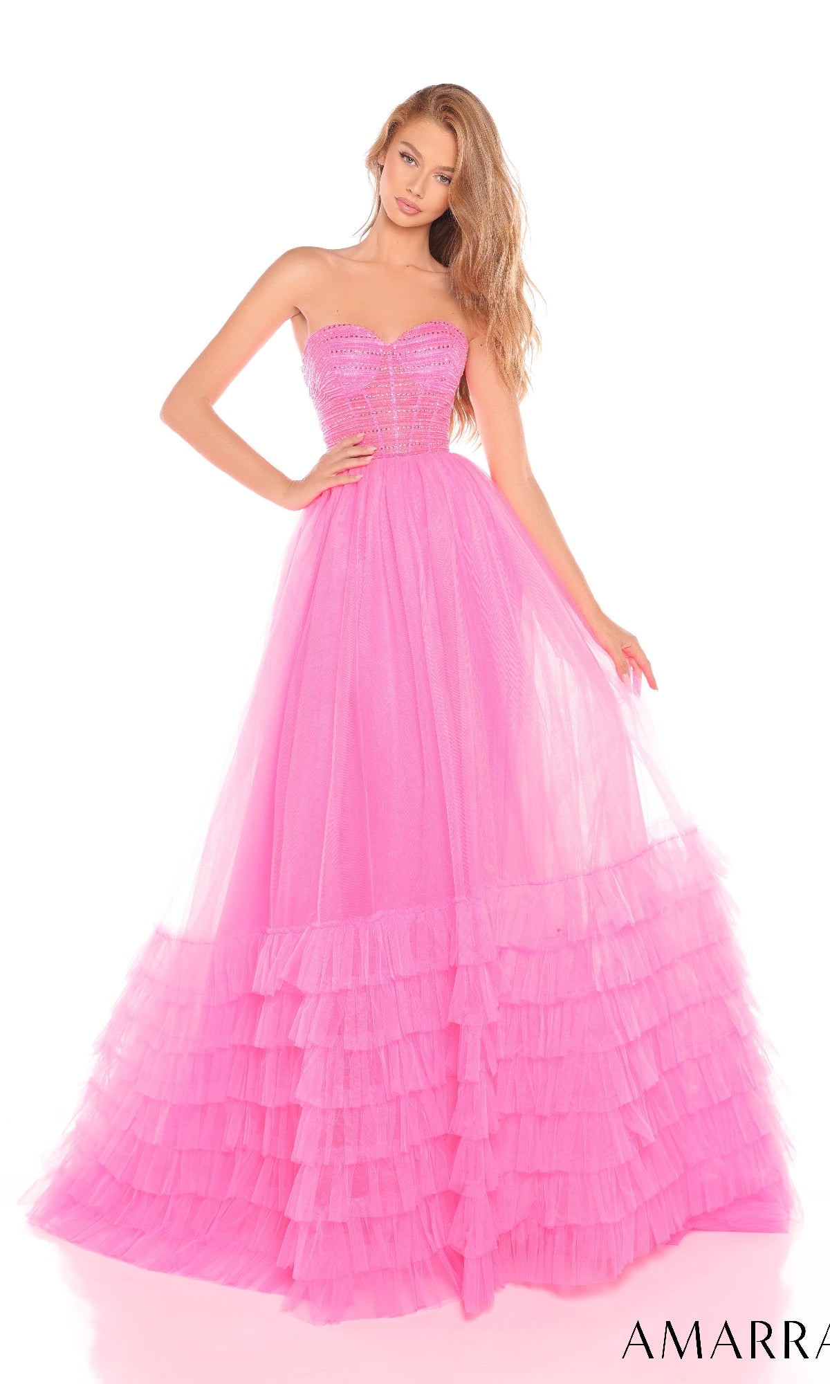 A woman poses against a white background in the Amarra 88277 Strapless Prom Ball Gown, featuring a sweetheart neckline and bright pink tulle a-line ruffled skirt.