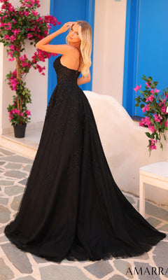 A woman in the Amarra 88416 long black strapless prom dress with a corset bodice stands outdoors by blue doors and potted flowering plants on a stone walkway.