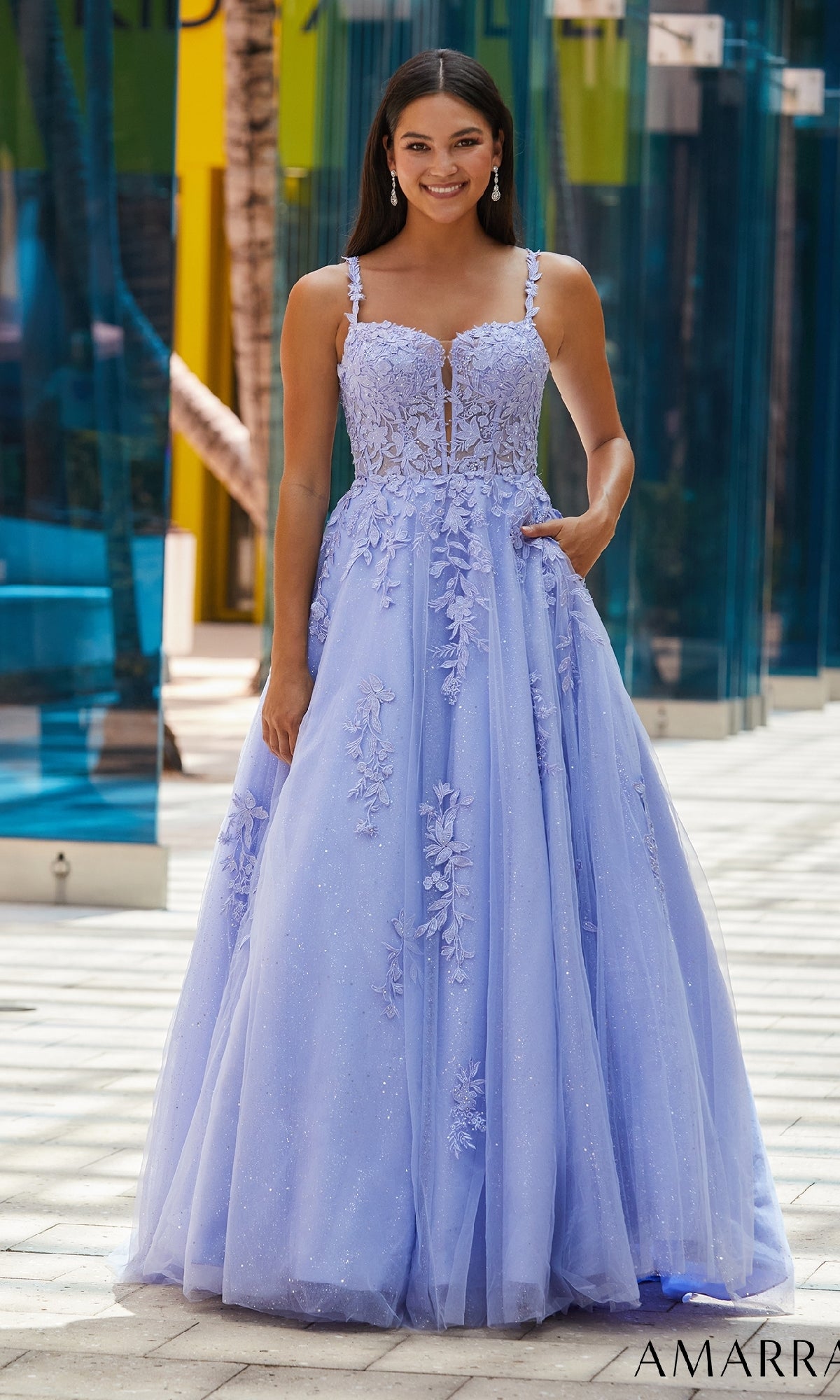 A woman stands outdoors on a tiled walkway wearing the Amarra 88590, a sleeveless floor-length lavender Floral Lace Prom Ball Gown with floral embellishments.