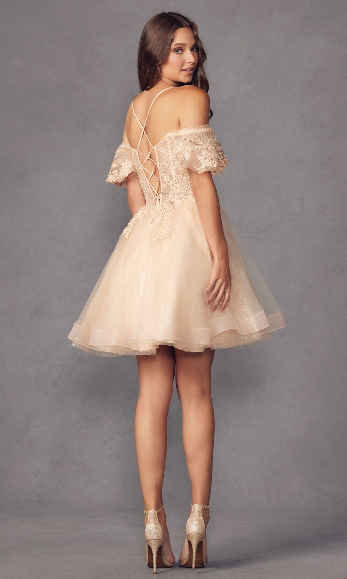 A woman wears the Puff-Sleeve Short A-Line Shimmer Prom Dress 886—an affordable, light beige, off-the-shoulder semi-formal dress with puffed sleeves and a lace-up back—posing in high heels against a plain studio background.