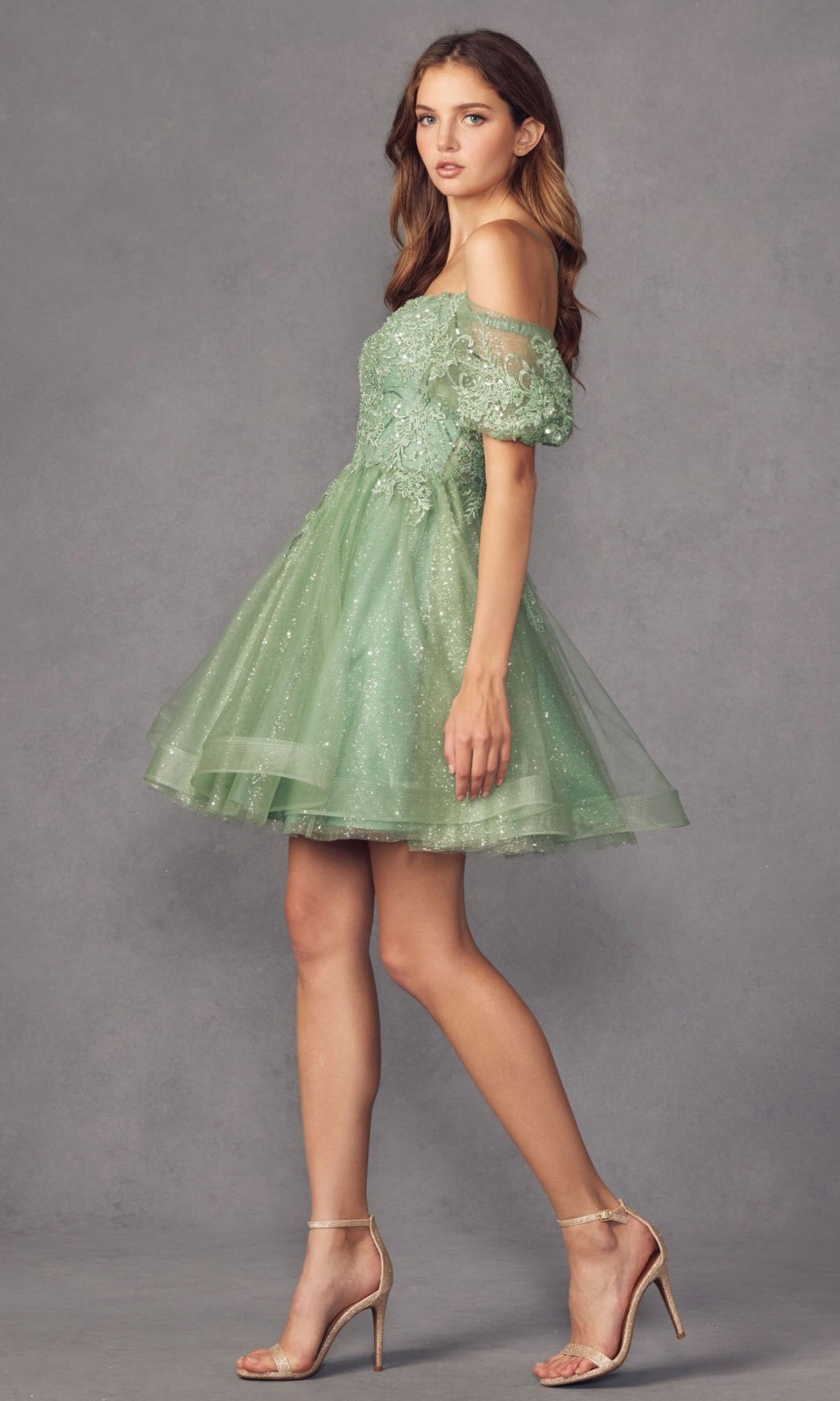 A woman stands against a gray background in the Puff-Sleeve Short A-Line Shimmer Prom Dress 886—a light green, off-the-shoulder, lace dress with sheer sleeves and sparkly accents—paired with nude high-heeled sandals.