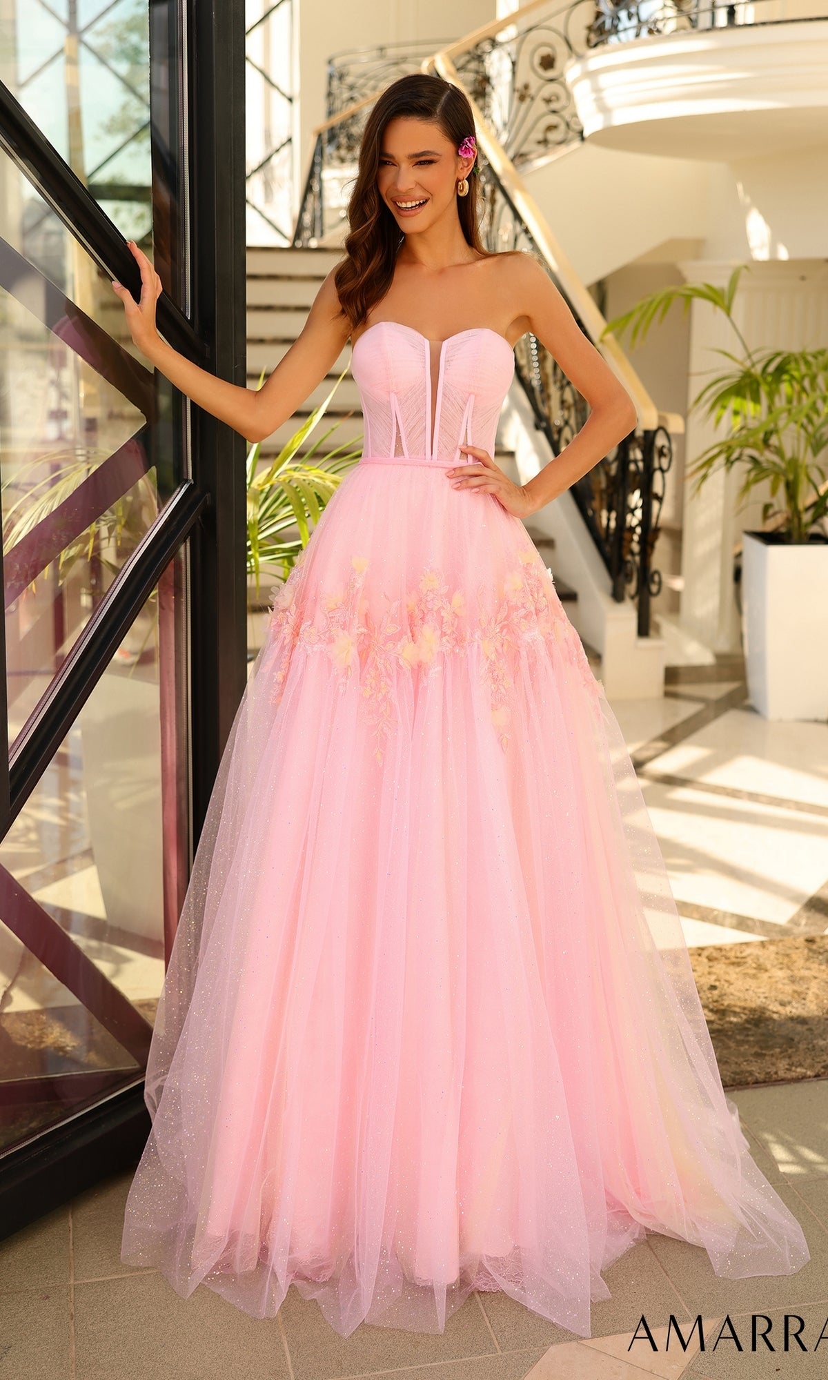 A woman wearing the Amarra Strapless Long Glitter Prom Ball Gown 88874 with floral details and a corset-style bodice stands indoors near stairs and glass doors, smiling with one hand on the doorframe.