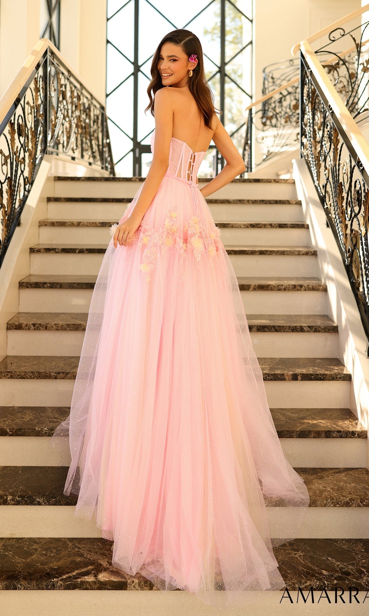 A woman in the Amarra Strapless Long Glitter Prom Ball Gown 88874, a floor-length pink tulle dress with a corset-style bodice and floral accents, stands on a staircase and looks over her shoulder.