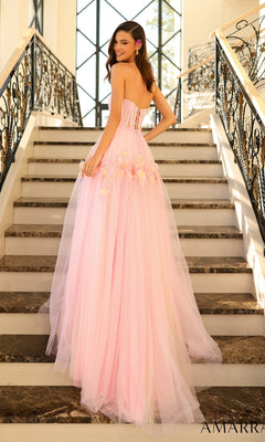A woman in the Amarra Strapless Long Glitter Prom Ball Gown 88874, a floor-length pink tulle dress with a corset-style bodice and floral accents, stands on a staircase and looks over her shoulder.