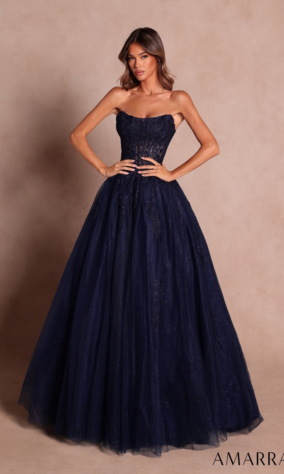 A woman stands against a neutral background wearing the Strapless Long Prom Ball Gown: Amarra 88913 in Navy, featuring a fitted, sheer lace corset-style bodice and a full tulle skirt.