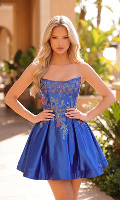 A woman stands outdoors with her hands on her hips, wearing the Amarra 88962 Strapless Fit-and-Flare Hoco Dress in royal blue, adorned with floral embroidery, and her long wavy blonde hair flowing over her shoulders.
