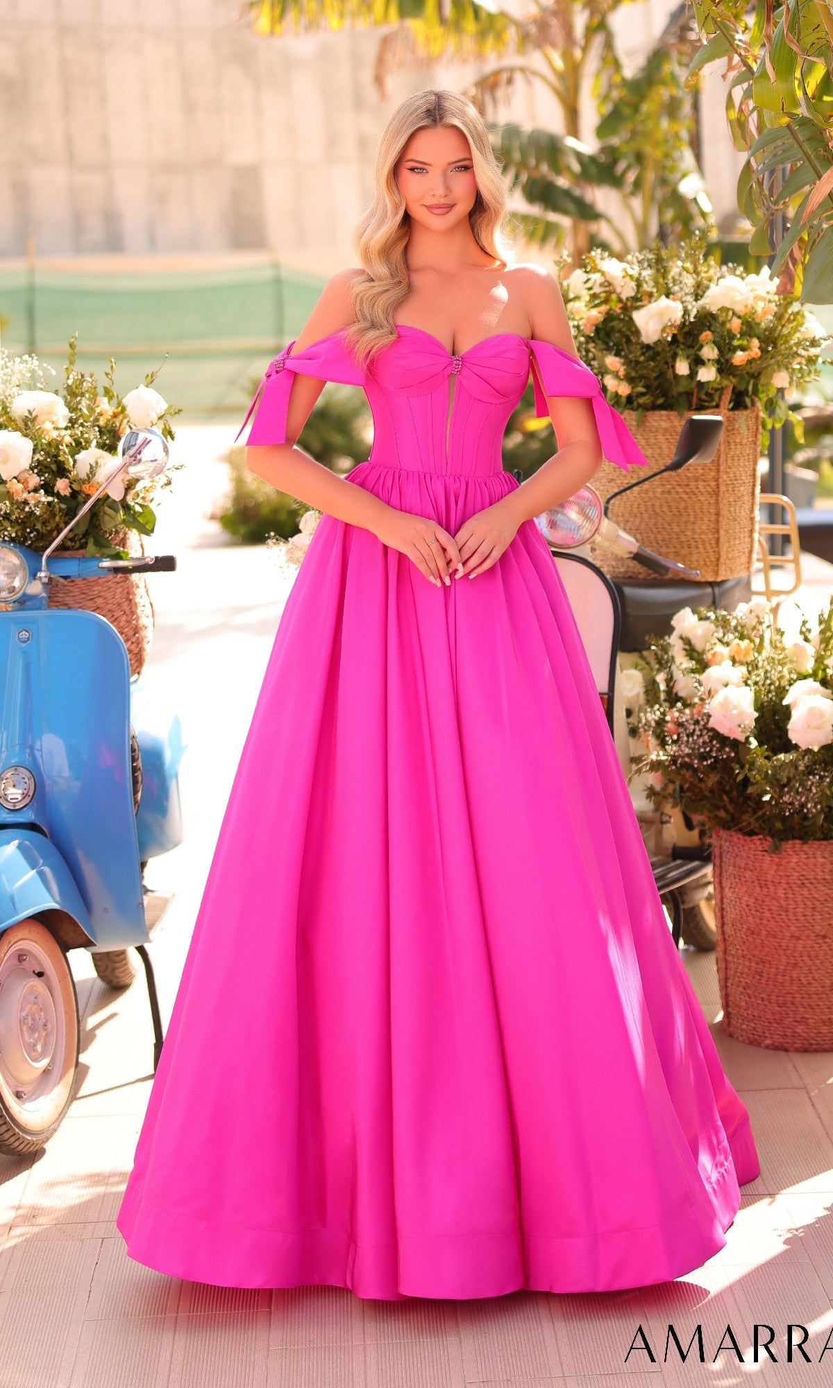 A woman stands outdoors surrounded by flowers and a blue scooter, wearing the Amarra 88995 Lace-Up Off-Shoulder Prom Ball Gown in bright pink.