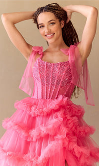Woman wearing a pink ball gown with ruffled layers and sheer shoulder ties against a beige background