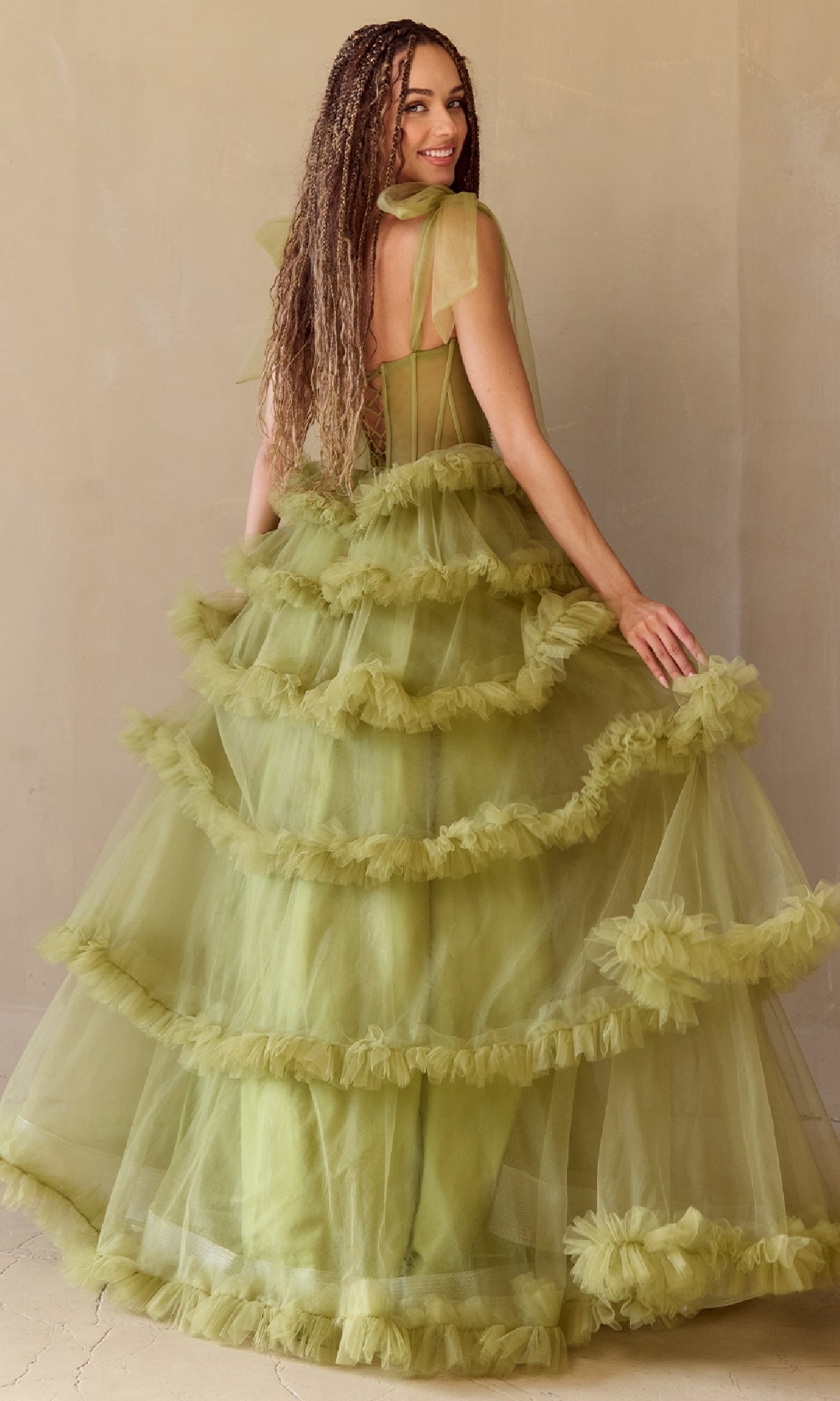Woman modeling the back of a green ball gown with a tiered skirt, in front of a beige background.