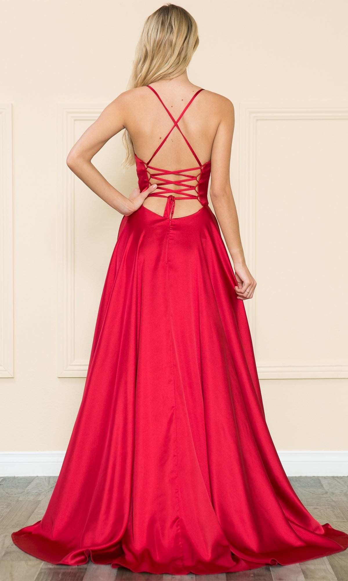 A woman with long blonde hair faces away from the camera indoors, wearing the Lace-Up Corset-Back Long A-Line Prom Dress 8910, a floor-length red satin gown featuring an open lace-up back.