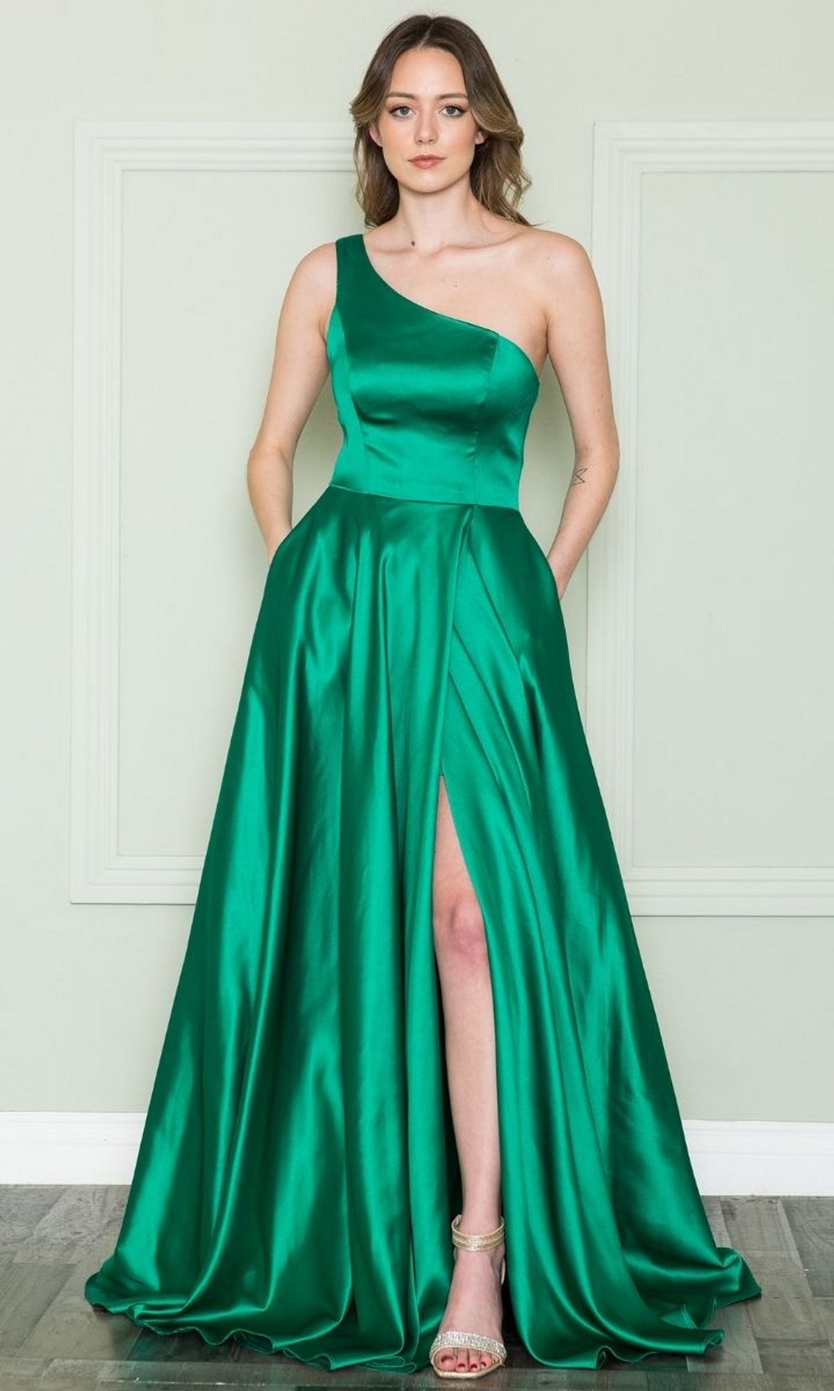 A woman wears the One-Shoulder Long Prom Dress with Pockets 8912 in emerald—an elegant evening gown featuring a thigh-high slit and side pockets, styled with strappy heels for a standout, budget-friendly look.