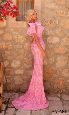 A woman in the Amarra 89175 long prom dress—a fitted, embellished pink one-shoulder gown with a large bow—stands against a stone wall beneath red flowers.