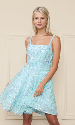 A woman in the Short A-Line Sequin Homecoming Dress 8930, featuring thin straps and light blue sequins, stands before a beige wall, softly smiling while holding the skirt with one hand.