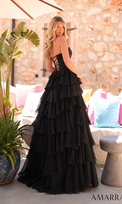 A woman stands outdoors near colorful cushions and a stone wall, wearing the Amarra 89311 long prom dress with a strapless corset bodice and layered black chiffon A-line skirt.