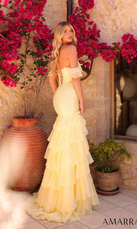 A woman wearing the Amarra 89345 long prom dress in light yellow, featuring off-the-shoulder tiered organza ruffles, stands outdoors in sunlight, surrounded by stone walls and vibrant pink flowers, facing away from the camera.