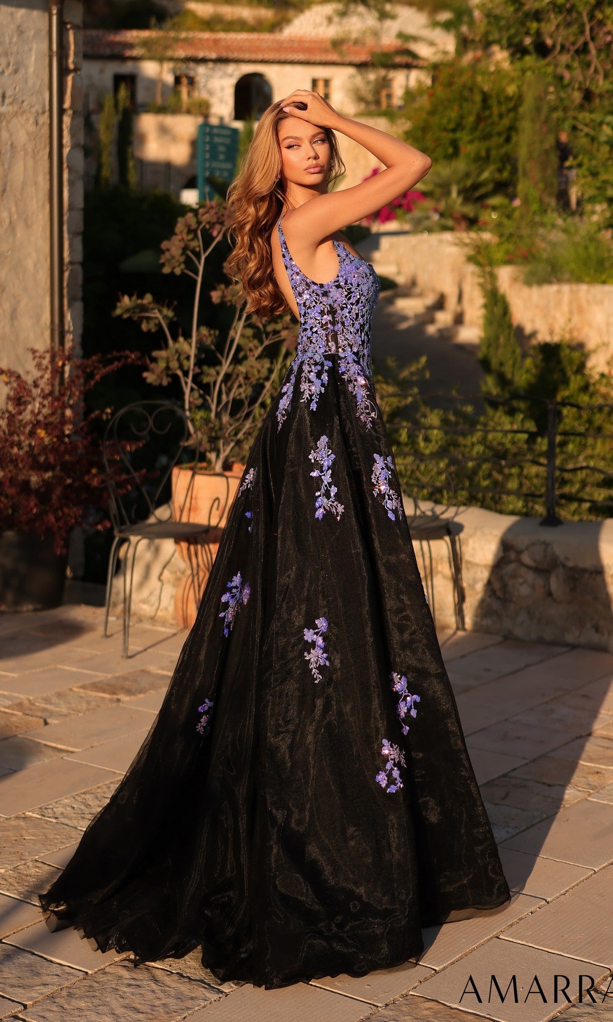 A woman poses outdoors in the Amarra 89373 long prom dress, featuring a sleeveless design, purple floral accents, and a fitted silhouette, as she stands on a stone patio surrounded by lush greenery.