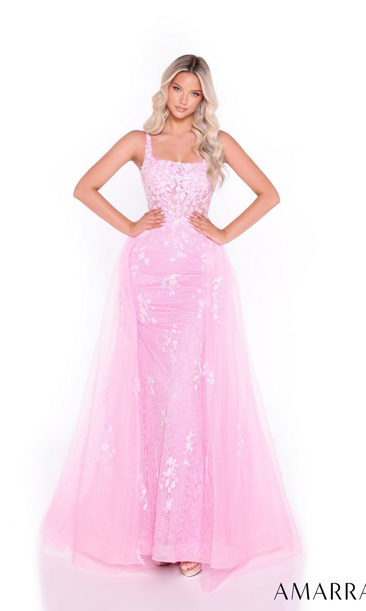 A woman stands with hands on hips against a white background, wearing the Amarra 89373 long prom dress—featuring a fitted silhouette, floor-length pink gown, and floral-embellished tulle overskirt with a sheer overlay.
