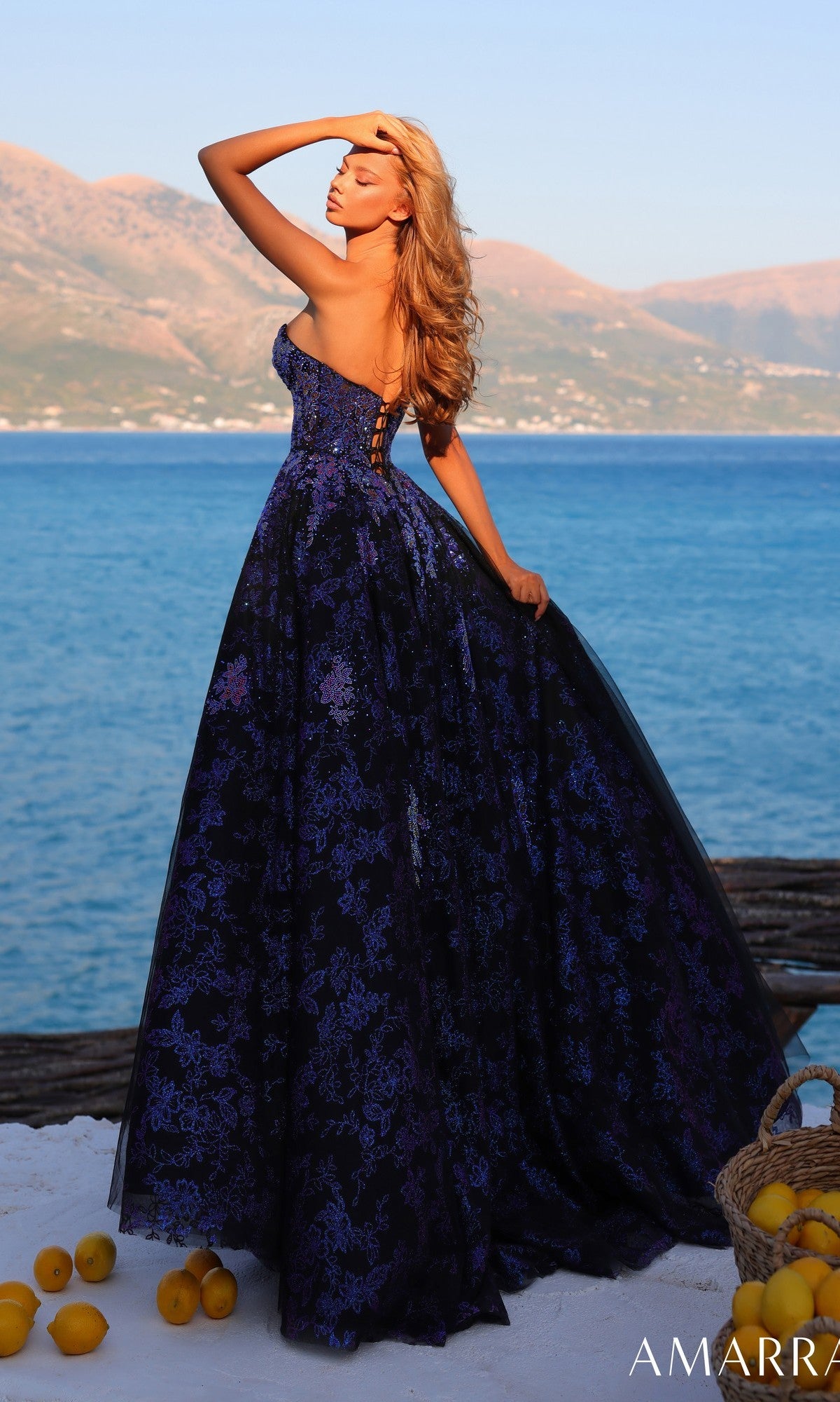 A woman poses by the sea and mountains with a basket of lemons, wearing the Amarra 89415 long prom dress featuring a strapless bodice and an A-line tulle skirt.