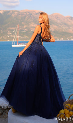 A woman in the Amarra 89429 Sheer-Corset Long Prom Dress, featuring a dark blue embroidered bodice, stands outdoors by the sea with mountains and a sailboat in the background.