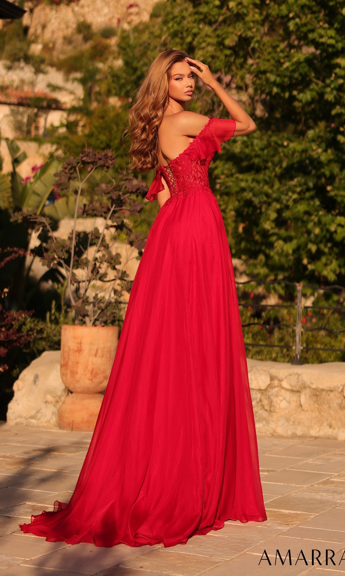 A woman stands outdoors in the Amarra 89481 long prom dress, featuring an off-shoulder design, flowing chiffon skirt, and delicate lace details, as she faces away and looks over her shoulder.