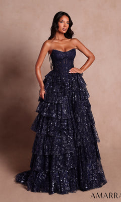 A woman stands against a plain beige background, wearing the Strapless Ruffled Prom Ball Gown: Amarra 89483 in navy. This elegant ball gown features a stunning tiered design with a delicate lace overlay, capturing the timeless sophistication of Amarra—perfect for making an unforgettable entrance.
