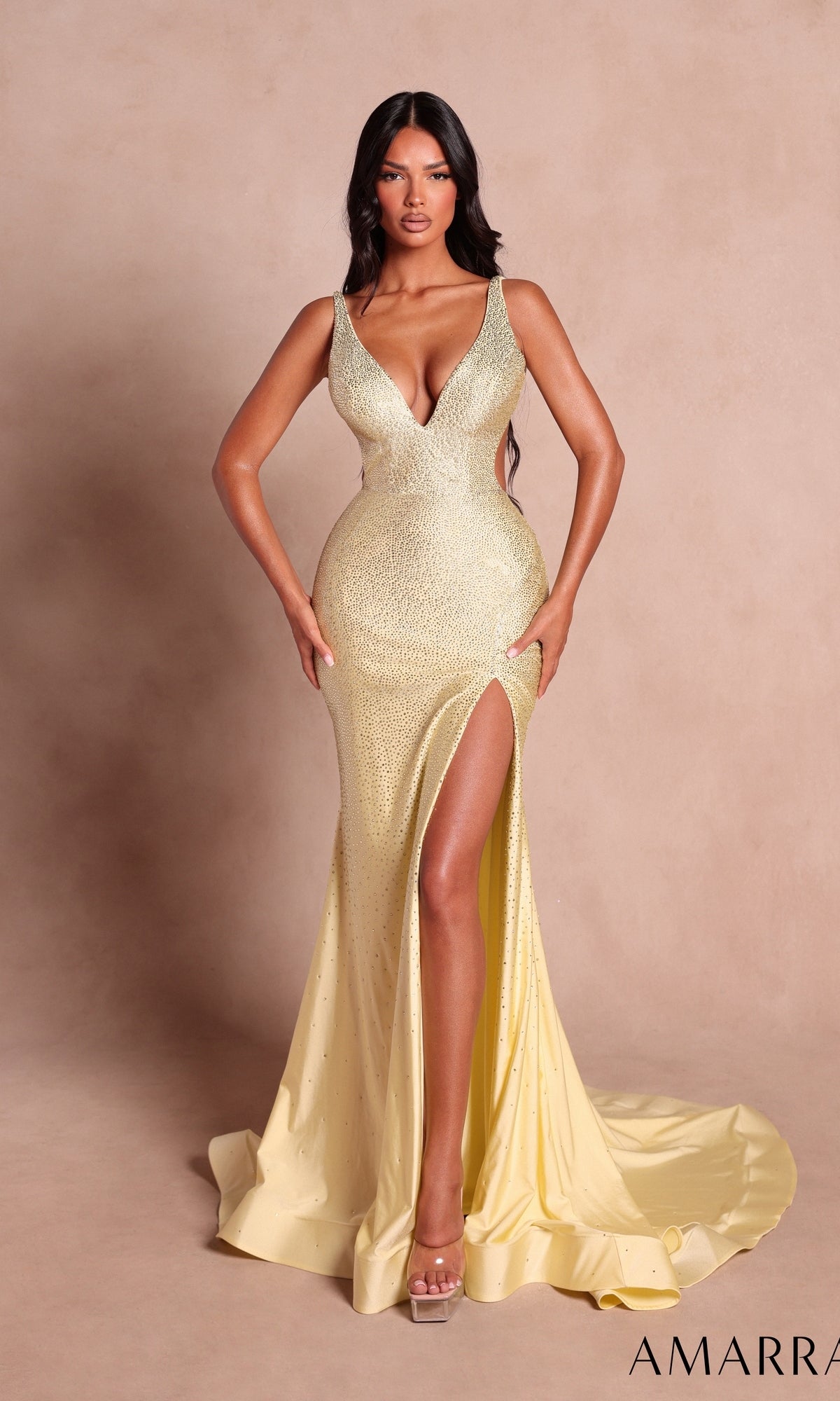 A woman wearing the Amarra 89503-1 long prom dress in light yellow, featuring sparkling embellishments, a deep V-neckline, fitted bodice, and a high leg slit, stands against a beige backdrop.