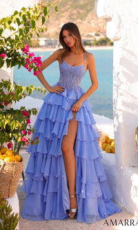 A woman wearing the Amarra 89507 long prom dress with an embellished corset bodice and tiered chiffon skirt stands by white walls, flowers, baskets of lemons, and a scenic backdrop of water and mountains.