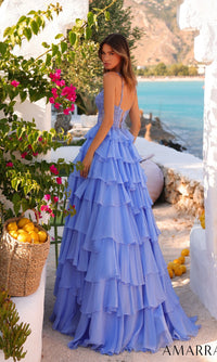 A woman wearing the Amarra 89507 long prom dress with an embellished corset bodice and tiered chiffon skirt stands outdoors by the sea, surrounded by white walls, greenery, and a basket of lemons.