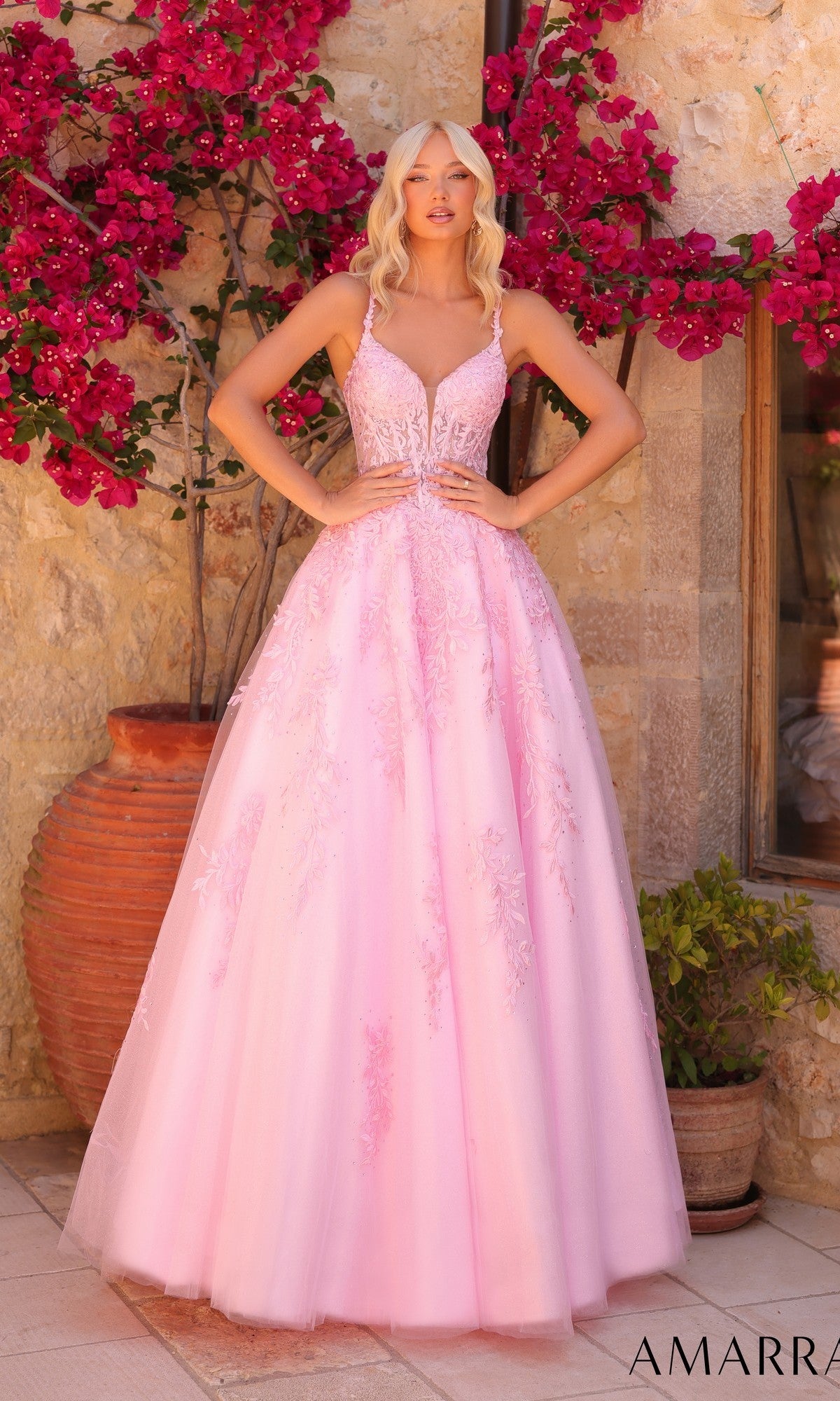 A woman stands outdoors by a stone wall and pink flowers, wearing the Amarra 89509 long prom dress with a lace corset bodice, floral embroidery, and a deep V neckline in light pink.