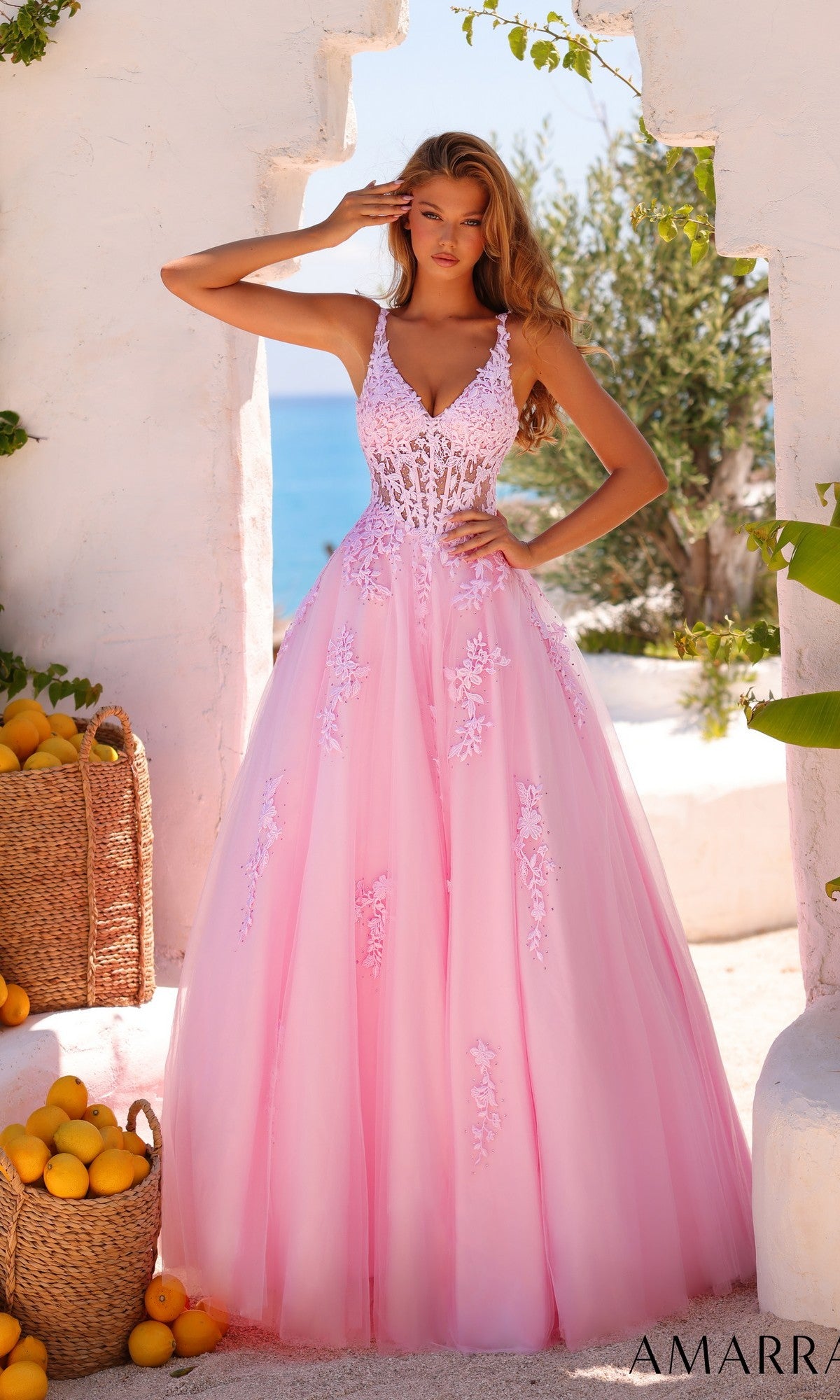 A woman in the Amarra 89523 Sheer-Waist Long A-Line Prom Dress in light pink, featuring floral lace embroidery, stands outdoors surrounded by white walls, baskets of lemons, and lush greenery.