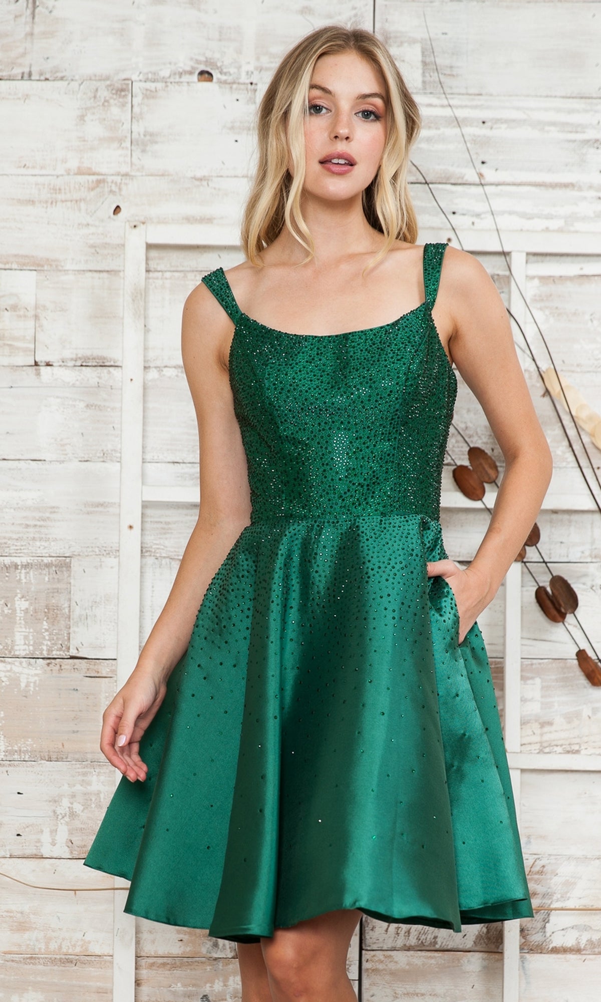 Woman wearing the Beaded Short A-Line Homecoming Dress 8958, featuring a sleeveless, fitted bodice and flared skirt, standing before a rustic wooden background.