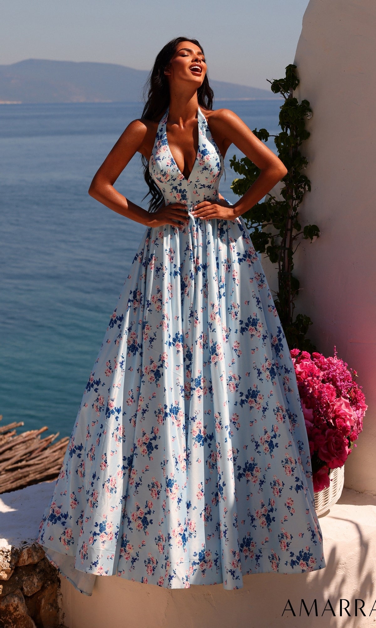 A woman wearing the Amarra 89616 long prom dress in blue/print, featuring a floral halter design and an A-line skirt, stands outdoors by the sea next to a white wall and a pot of pink flowers under clear daylight.