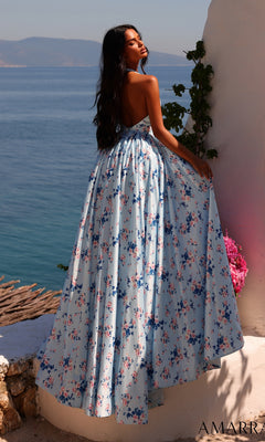 A woman wearing the Long Prom Dress: Amarra 89616, a blue floral-printed halter gown, stands by a white wall overlooking the sea, with pink flowers and mountains visible in the background.