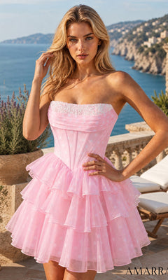 A woman stands outdoors on a terrace with coastal scenery and hills in the background, wearing the Amarra 89623 Short Homecoming Dress in Pink/White—a strapless, tiered fit-and-flare style.