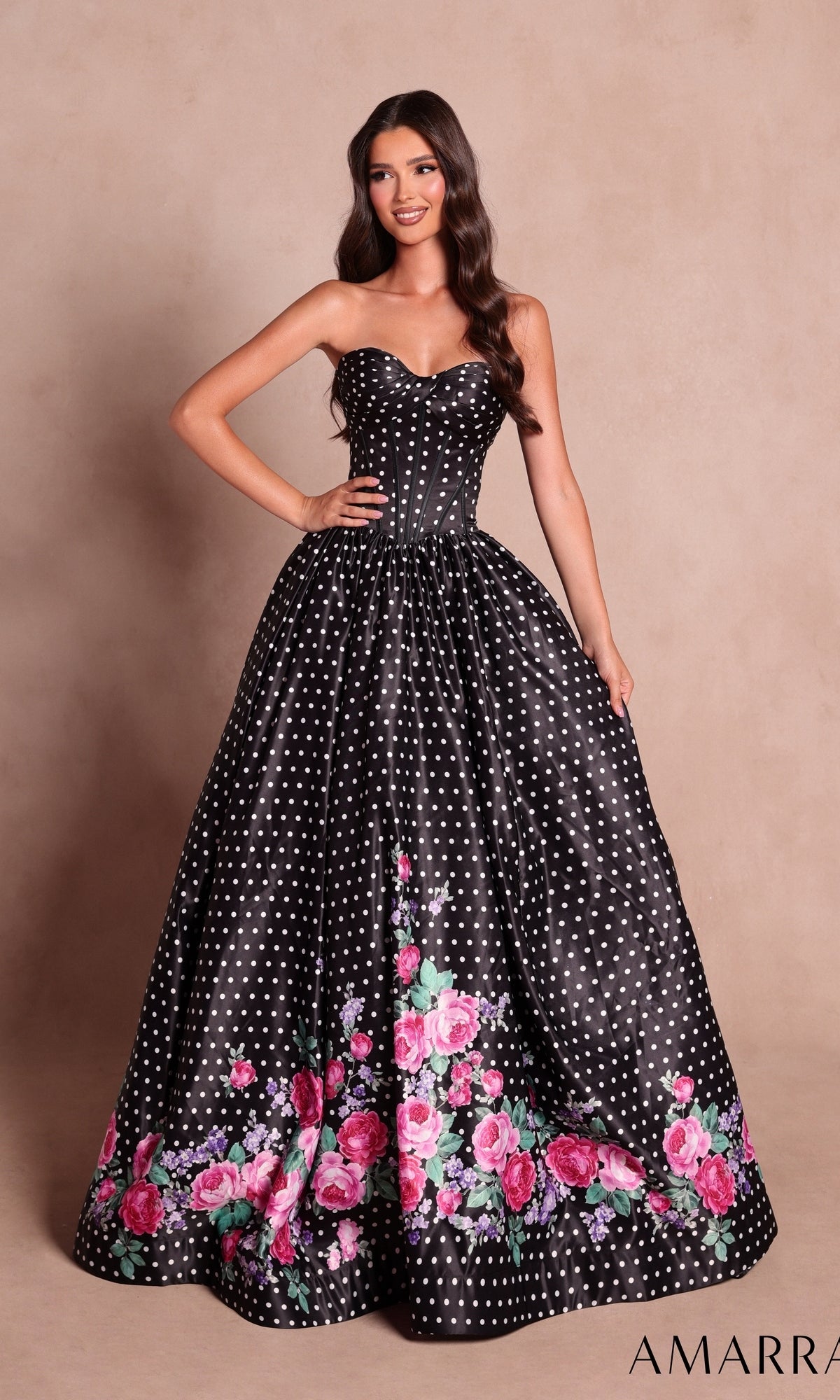 A woman wearing the Amarra 89624 long prom dress in Black/Print, a strapless ball gown with a corset bodice, decorated with white polka dots and pink floral print along the hem, stands against a neutral background.