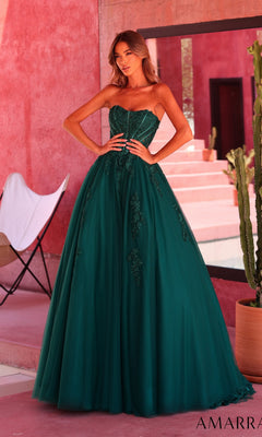 A woman wearing the Amarra 89626 long prom dress in emerald, featuring a strapless design with lace details and a subtle sweetheart neckline, stands indoors near pink stucco walls, potted plants, and a white chair.