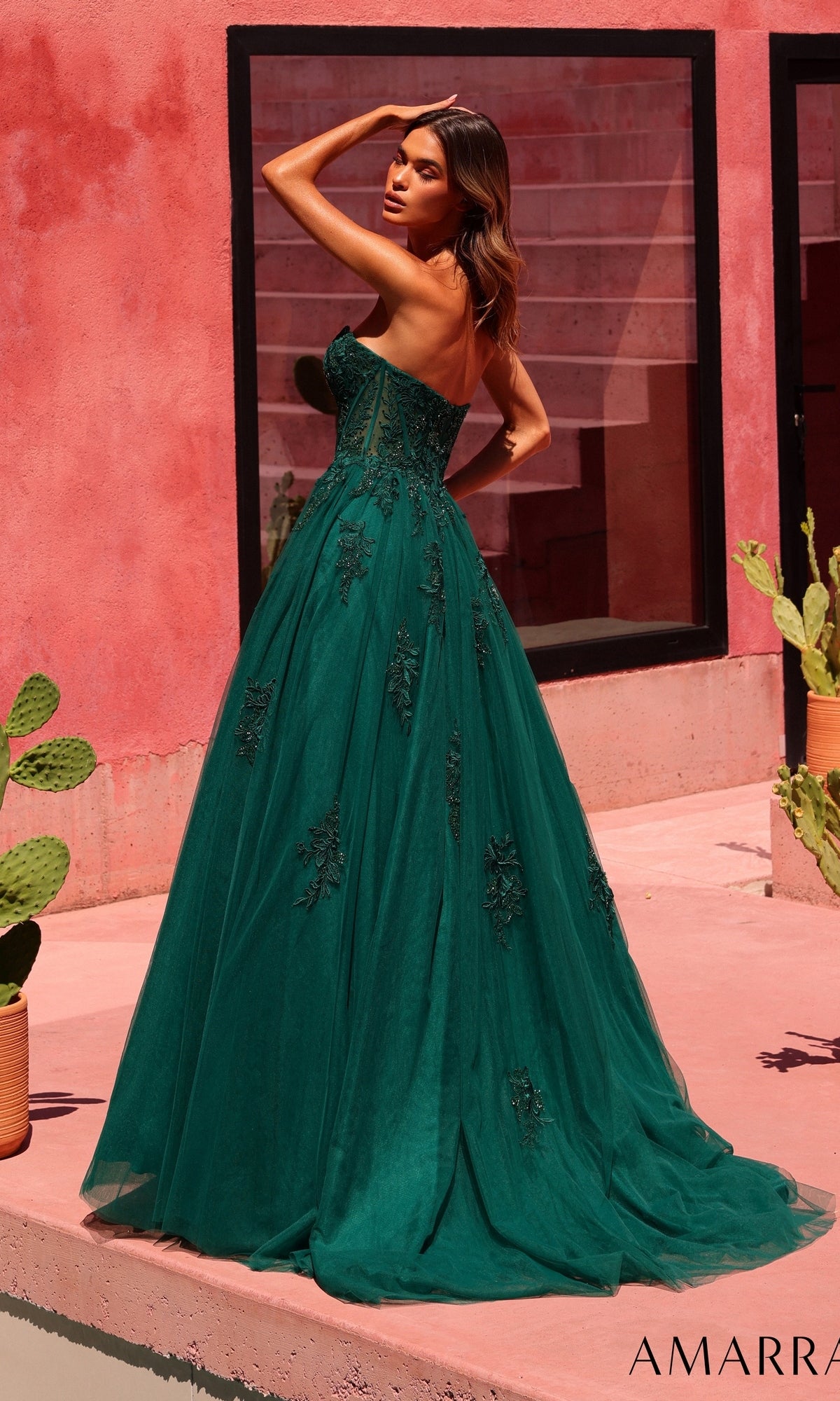 A woman stands outdoors on a pink patio, facing away and looking over her shoulder, wearing the Amarra 89627 long prom dress—a strapless emerald green gown adorned with beaded floral lace embroidery.