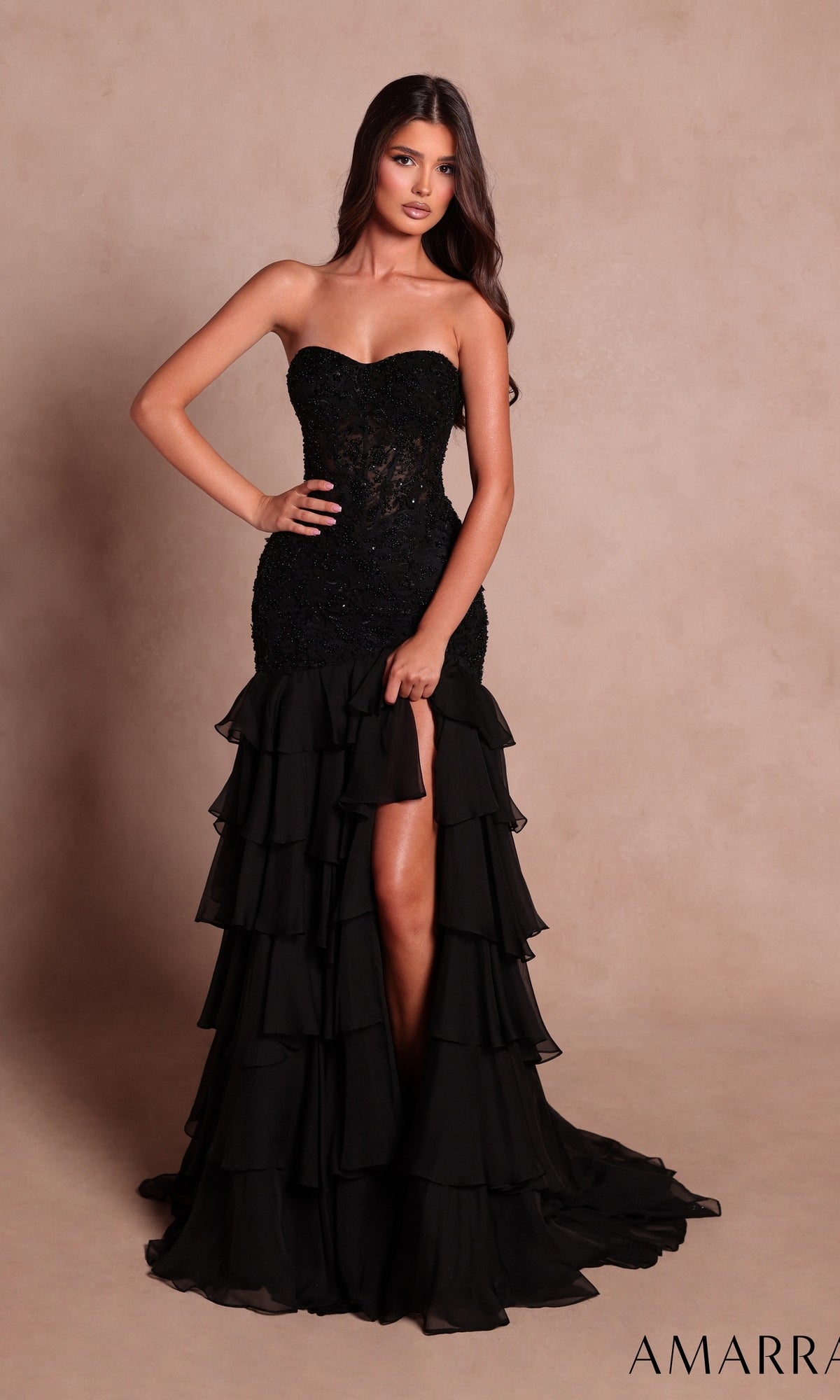 A woman wearing the Amarra 89631 long prom dress in black, featuring a strapless fitted bodice, tiered ruffles along the layered skirt, and a front slit, stands against a plain beige background.