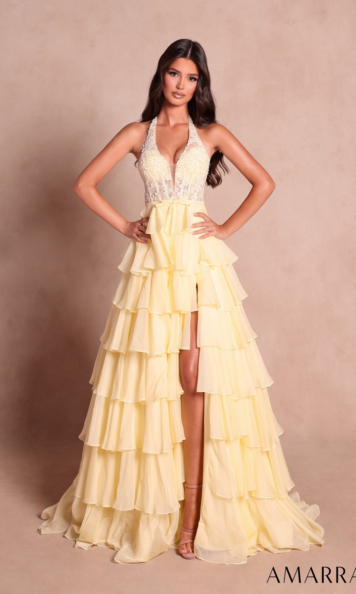 A woman stands against a plain beige background wearing the Amarra 89633 long prom dress in light yellow, which features a halter neckline, beaded bodice, and a tiered ruffle skirt.