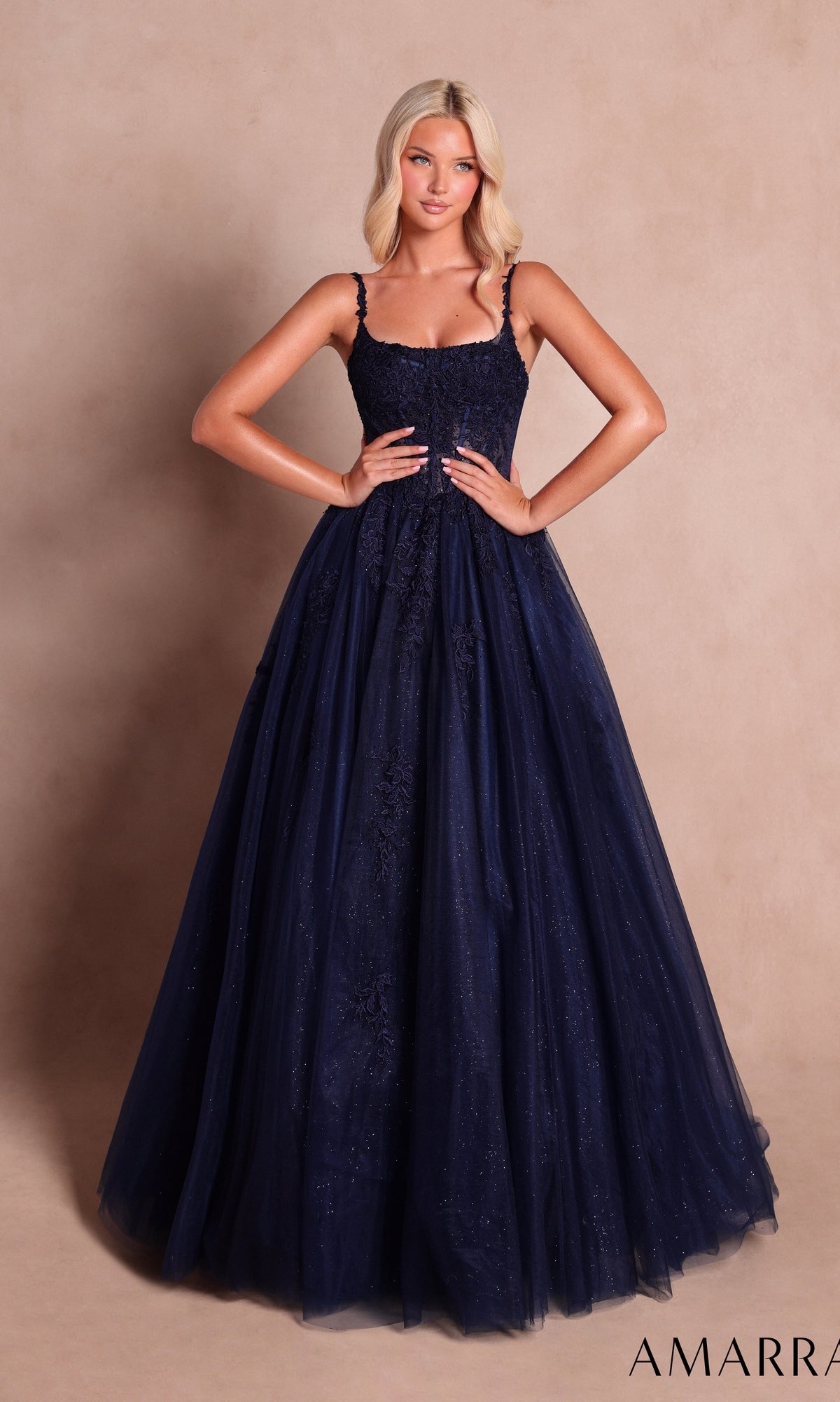 A woman wearing the Long Prom Dress: Amarra 89644 in navy, a sleeveless, floor-length gown featuring a sweetheart neckline, full skirt embellished with subtle sparkle and delicate floral lace appliqués, stands against a plain beige background.