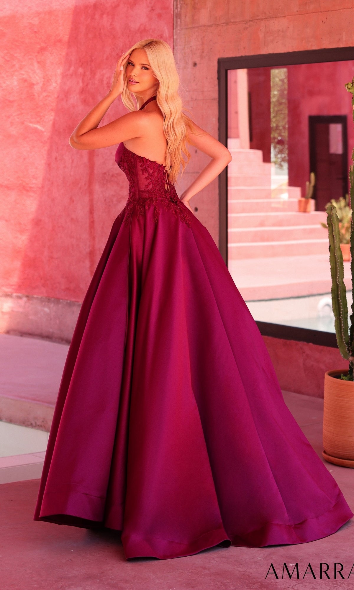 Wearing the Amarra 89646 Long Prom Dress, a woman in a strapless, floor-length magenta ball-gown stands indoors near a staircase and cactus, facing away with one hand touching her hair.