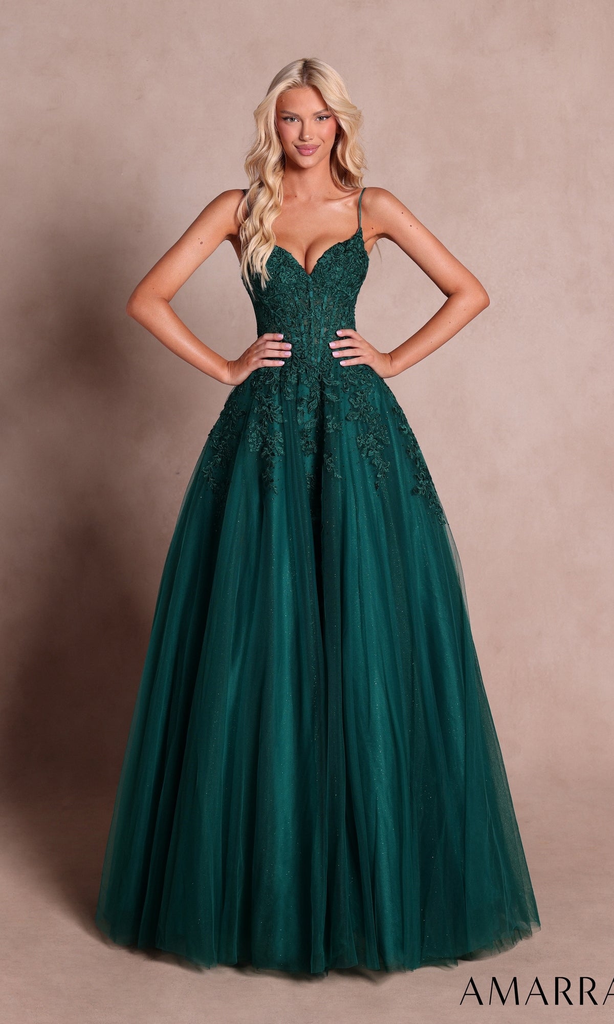 A woman wearing the Amarra 89682 Long Prom Dress in emerald, featuring a lace bodice and a tulle ball-gown skirt, stands against a beige background with her hands on her hips.