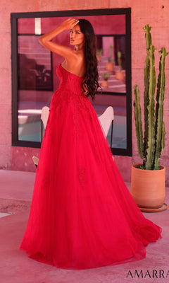 A woman wearing the Amarra 89684 Long Prom Dress, a strapless red lace gown with a flowing tulle A-line skirt that enhances her silhouette, stands indoors facing away with one hand raised to her head. Behind her are a tall potted cactus and a large window.
