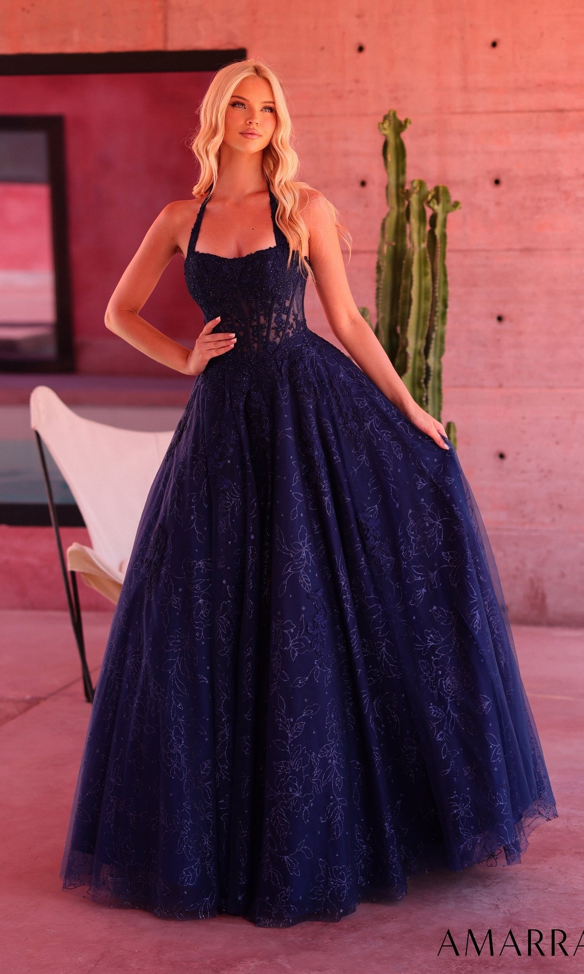 A woman stands indoors near a cactus wearing the Amarra 89685 prom ball gown in navy, a floor-length dress with a halter neckline and lace details. Elegant glitter floral print accents add a touch of sparkle to this sophisticated look.