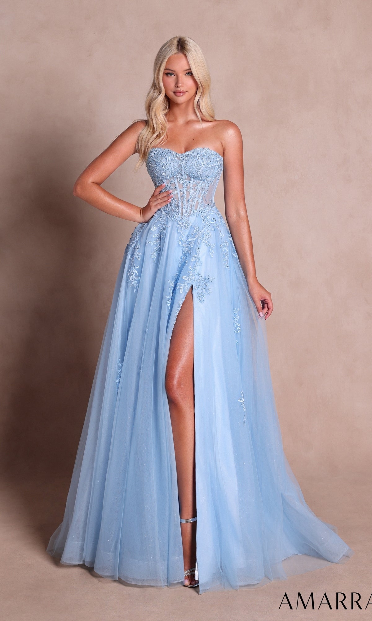 A woman poses in the Amarra 89689 long prom dress—a light blue gown featuring floral embroidery, a sheer bodice, and a thigh-high slit—standing against a beige background.