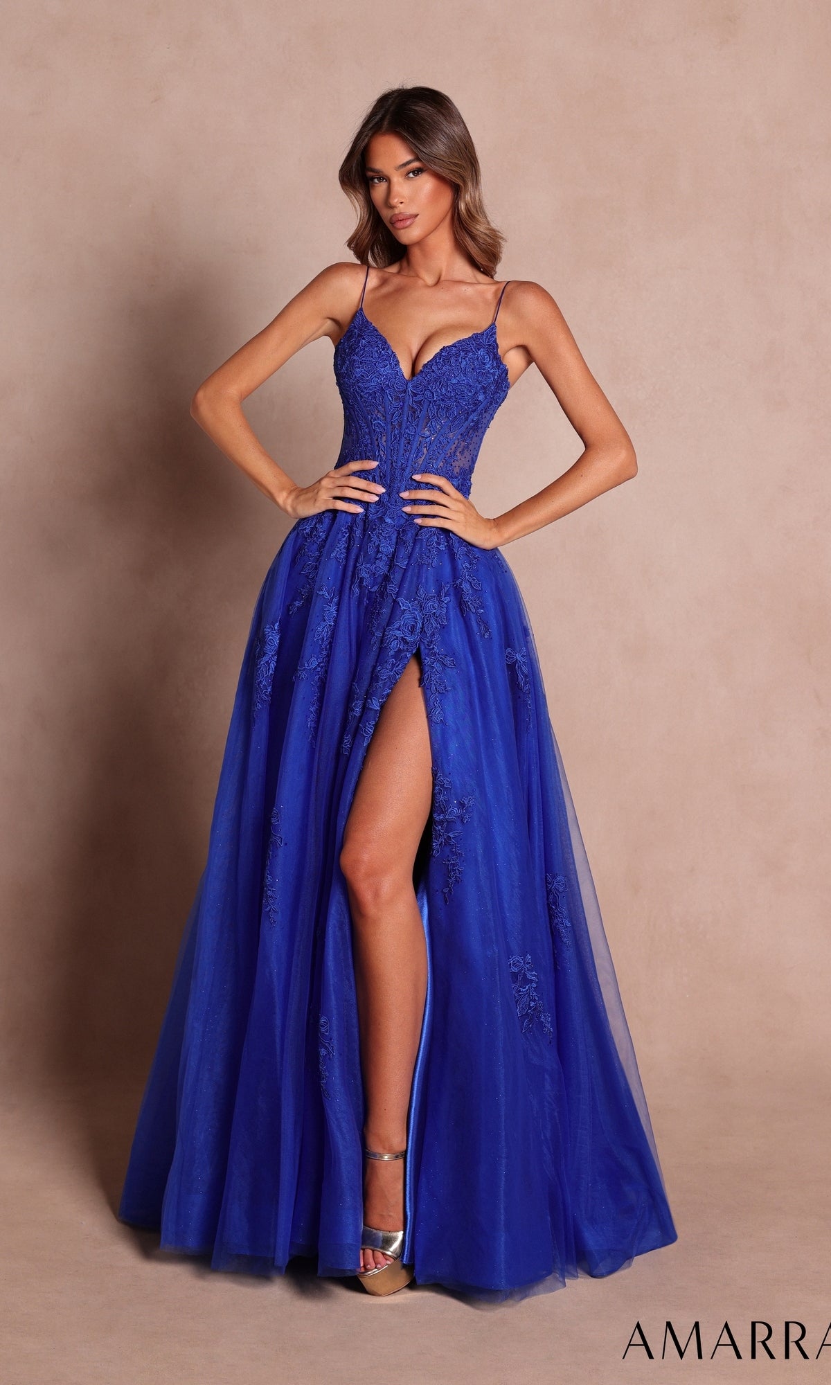 A woman models the Amarra 89693 long prom dress in royal blue—an elegant sleeveless evening gown adorned with floral lace appliqué, featuring a deep V-neckline, boned-corset bodice, and a high slit—posed against a plain beige background.