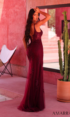 A woman wearing the Amarra 89695 long prom dress, featuring a sleeveless burgundy lace design with a fitted corset bodice, stands sideways and looks over her shoulder next to a potted cactus in a modern, pink-toned setting.